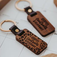 Dark stained wood keychain close up showing detail of the laser engraved "Coffe, Contracts and Closing" along with a cheetah print around the edge of the keychain. Gold colored keyring is secured to the wood keychain by a vegan leather strap secrured with a gold colored metal stud. 