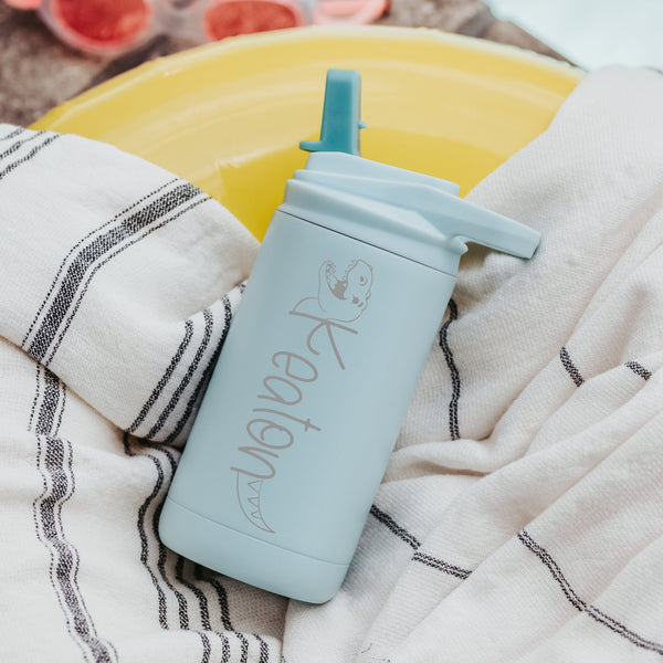 Mint color 12-ounce stainless steel, flip-top straw tumbler. The tumbler is personalized with a dinosaur that its body is the name of the child. The tumbler is nestled on a beach towel. 