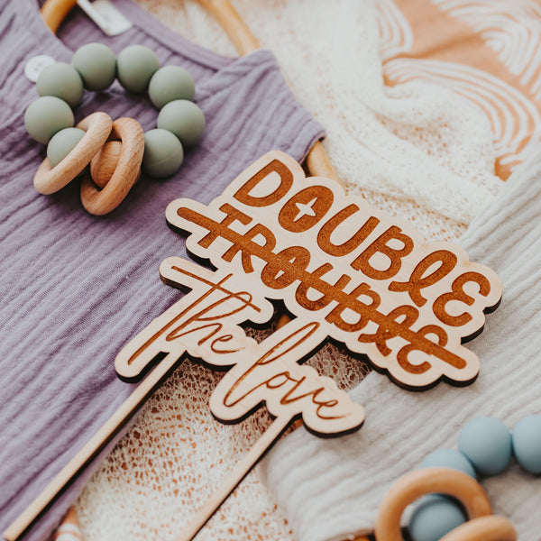 Double The Trouble Double the Love Cake topper on pastel colored baby clothes by Legacy and Light