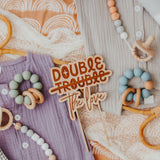 Double The Trouble Double the Love Cake topper on pastel colored baby clothes with baby toys by Legacy and Light