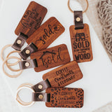 Dark-stained wood keychains with celebration phrases for buying a house. Each keychain has a different inscription: "I make dreams come true", "Sold", "Coffee, Contracts and Closings", "Sold is my favorite four-letter word". Each keychain's metal keyring is secured to the wood by a small strip of vegan leather and a metal stud.