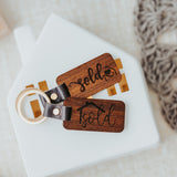 Two dark stained wood keychains both stating "Sold" in different formats on a white house background. 
