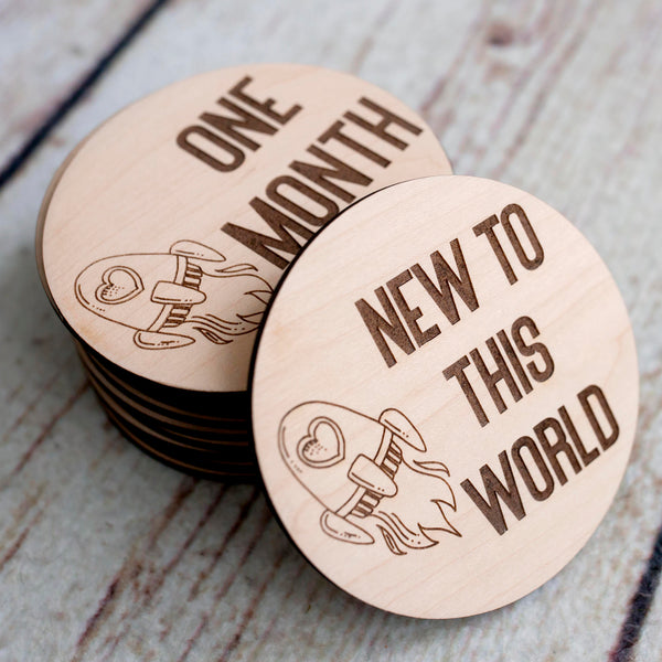 Round Birch wood baby milestone discs. Laser engraved with a rocket ship design with the phrase "new to this world" and the One month through 12 months.