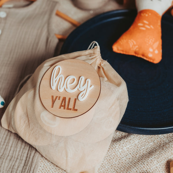 A wooden milestone card with the greeting 'hey y'all' in elegant white script, resting on a muslin drawstring bag. The bag contains a set of monthly milestone cards for a newborn baby, designed to capture and celebrate each month of the baby's first year. The card and bag are part of a cozy nursery setting, with a navy blue hat and an orange patterned pillow in the background, creating a warm and welcoming atmosphere.