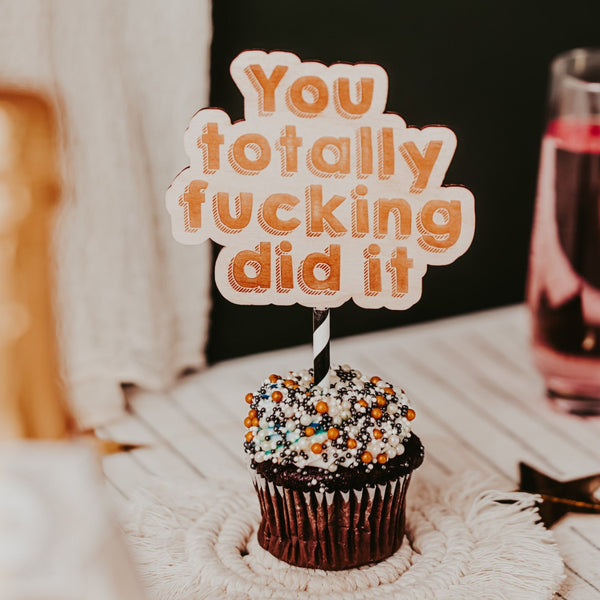 A cake topper with the writing in orange "YOU TOTALLY F*$&ing DID IT"